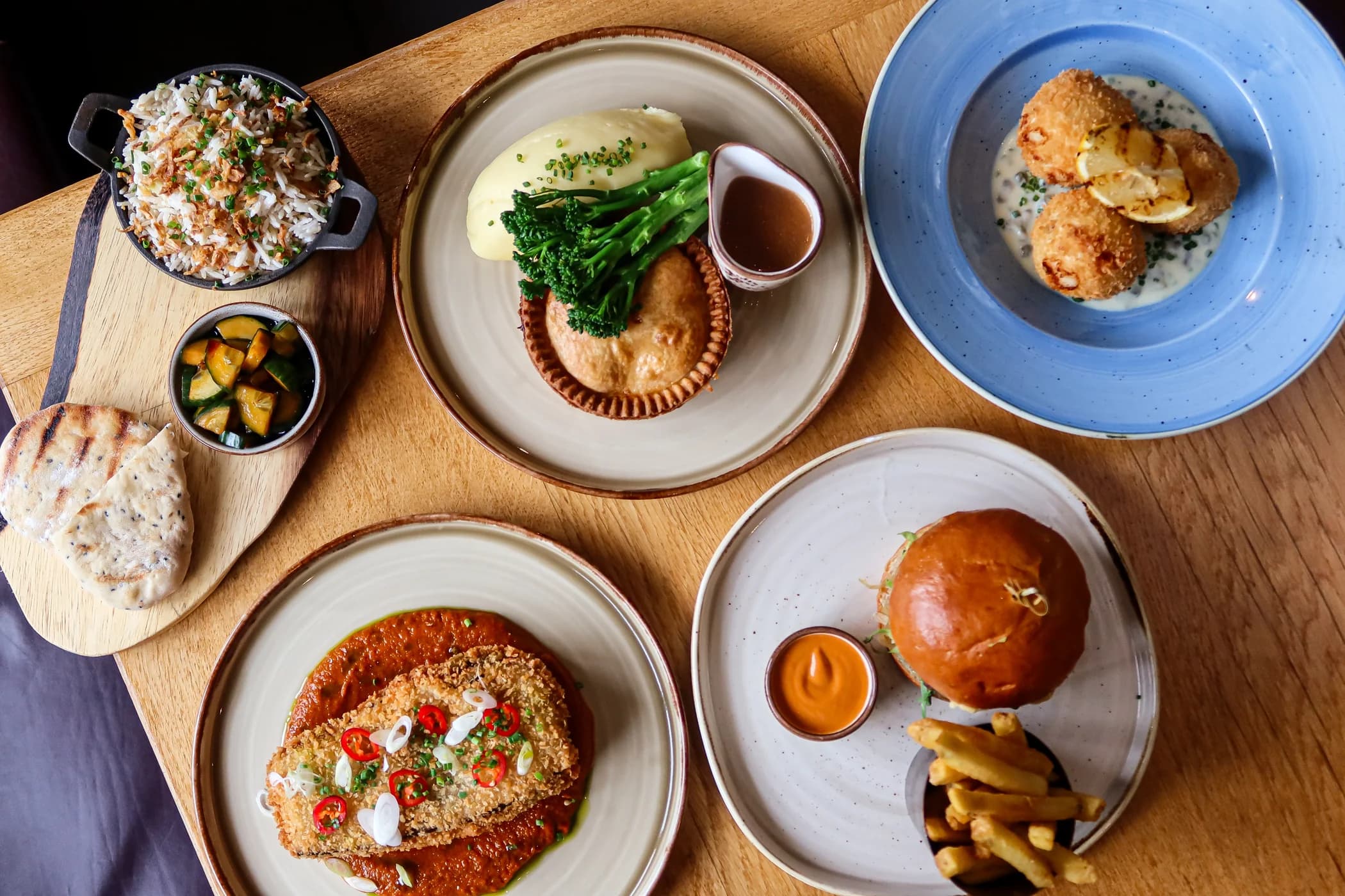 A spread of dishes at The Piccadilly Inn seen from above: a pie with mash, tenderstem broccoli and gravy, a burger and fries, a breaded fillet on a red sauce, croquettes on a blue plate, and rice, flatbreads and sides on a wooden board